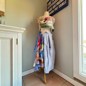 Könnte beinhalten: Eine Schaufensterpuppe mit einem floralen Oberteil und verschiedenen Hemden und Schals. Die Schaufensterpuppe steht in einer Ecke, mit einem Schild mit der Aufschrift "THE FONDEST MEMORIES ARE MADE GATHERED ROUND THE TABLE". Der Raum hat eine hellgrüne Wand und einen weißen Schrank.