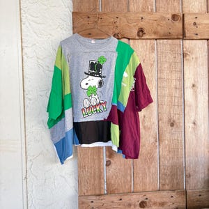 Könnte beinhalten: Ein Patchwork-T-Shirt mit Snoopy, der einen Zylinder und ein Kleeblatt trägt, darunter das Wort „LUCKY“. Das Shirt kombiniert graue, grüne, blaue und bordeauxfarbene Stoffbahnen. Es hängt an einem Metallbügel.