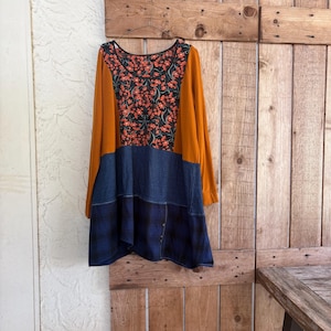 Puede incluir: Una túnica de manga larga con un diseño de bloques de color, mangas color amarillo mostaza, un panel central con estampado floral en negro y rojo, una sección media en azul sólido y un dobladillo inferior con patrón de cuadros azul oscuro y negro.