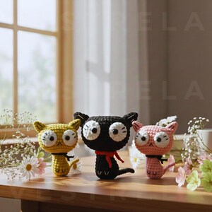 May include: Three handmade crochet cat figurines in yellow, black, and pink. Each cat has large white eyes, small ears, and a tail. The black cat wears a red scarf, and the yellow cat has a black collar.