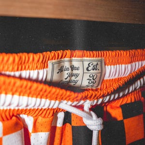 May include: Orange and black checkered basketball shorts with a white drawstring. The shorts have a white label that says "All in One Jersey Company Est. 2022".