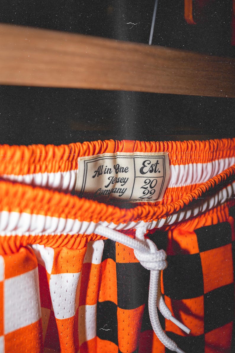 May include: Orange and black checkered basketball shorts with a white drawstring. The shorts have a white label that says "All in One Jersey Company Est. 2022".