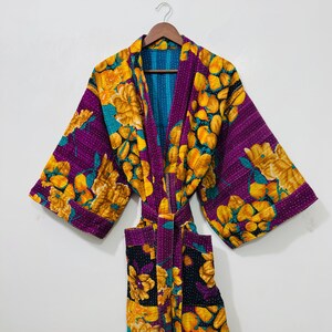 Może przedstawiać: Żywy, dwustronny szlafrok w stylu kimono z kwiatowym wzorem. Szlafrok ma fioletową podstawę z żółtymi i turkusowymi kwiatowymi wzorami. Wnętrze jest turkusowe, a szlafrok jest zabezpieczony pasującym paskiem. Szlafrok wisi na drewnianym wieszaku.