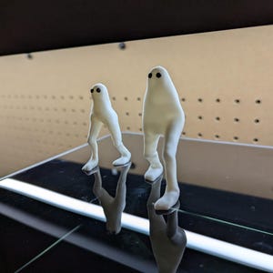 Peut inclure: Deux figurines blanches en forme de fantômes avec des yeux en points noirs se tiennent sur une surface réfléchissante. L'une est plus petite, avec des jambes visibles, tandis que l'autre est plus grande et plus arrondie. L'arrière-plan est un panneau perforé brun clair.