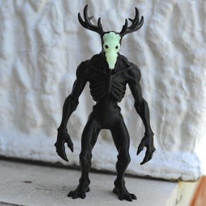 以下が含まれることがあります： 鹿の頭蓋骨と角を持つ生き物の黒い置物。頭蓋骨は淡い緑色です。生き物は骨格の胸と筋肉質な腕と脚を持っています。置物は灰色の表面に立っています。