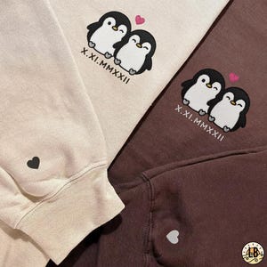 Könnte beinhalten: Drei Sweatshirts in Beige und Braun, jedes mit einem Pinguin-Paar-Design, einem rosa Herz und dem Datum X.XI.MMXXII. Ein Sweatshirt hat ein schwarzes Herz auf dem Ärmel, ein anderes ein weißes Herz.