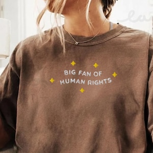 Pode incluir: Uma camiseta marrom de manga curta com o texto "BIG FAN OF HUMAN RIGHTS" em branco, com detalhes de estrelas amarelas. A camiseta é usada por uma pessoa segurando uma caneca branca.