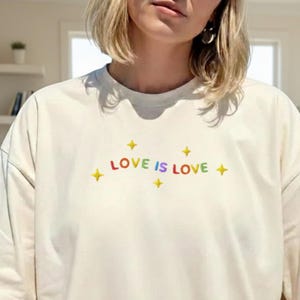 Könnte beinhalten: Cremefarbenes T-Shirt mit dem Text "LOVE IS LOVE" in Regenbogenfarben und gelben Sternakzenten. Das Shirt hat eine lockere Passform und kurze Ärmel. Das Design ist auf der Brust zentriert.