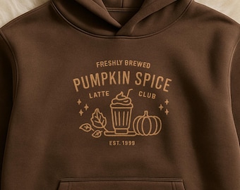 May include: A brown hooded sweatshirt with a front pocket. The sweatshirt features a graphic that reads "Freshly Brewed Pumpkin Spice Latte Club" with a coffee cup, pumpkin, and leaves. The text "Est. 1999" is below the graphic.