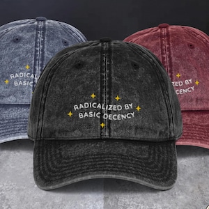 May include: Three baseball caps in acid-washed blue, black, and maroon. Each cap features the embroidered white text "RADICALIZED BY BASIC DECENCY" centred on the front, accented by small yellow four-pointed star icons surrounding the lettering.