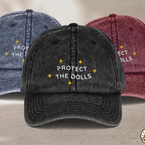Peut inclure: Trois casquettes de baseball en tissu délavé bleu, noir et bordeaux. Chaque casquette présente un texte blanc brodé indiquant PROTECT THE DOLLS, agrémenté de petites étoiles jaunes à quatre branches cousues autour du lettrage sur le devant.