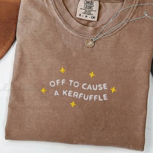 Könnte beinhalten: Espressofarbenes T-Shirt mit dem gestickten Schriftzug "OFF TO CAUSE A KERFUFFLE" und gelben Sternen. Eine silberne Halskette mit Anhänger liegt auf dem Shirt. Das Shirt ist von der Marke Comfort Colors, Größe S-P-CH.