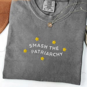 Puede incluir: Una camiseta gris Comfort Colors con la frase bordada "SMASH THE PATRIARCHY" en blanco, acentuada con estrellas amarillas. También se ven un collar plateado con un colgante y un sombrero marrón.