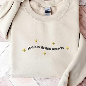 Könnte beinhalten: Ein cremefarbenes Sweatshirt mit Rundhalsausschnitt und dem aufgestickten schwarzen Schriftzug "MAUSIS GEGEN RECHTS" mittig auf der Brust, umgeben von fünf kleinen, gelben, vierzackigen Sternsymbolen in einer geschwungenen Anordnung.