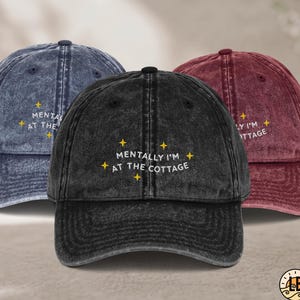 Könnte beinhalten: Drei Baseballkappen in Blau, Schwarz und Kastanienbraun mit verwaschenem Vintage-Finish. Jede Kappe zeigt den gestickten weißen Schriftzug "MENTALLY I'M AT THE COTTAGE", umgeben von kleinen gelben Sternsymbolen auf der Vorderseite.
