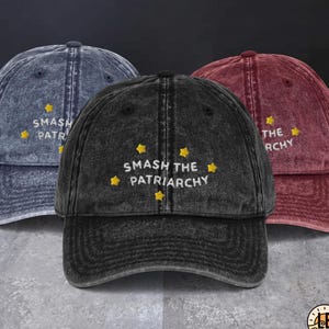 May include: Three baseball caps in acid-wash blue, black, and maroon. Each cap features embroidered yellow stars and the white text SMASH THE PATRIARCHY. The hats have a curved brim, a six-panel construction, and a button on the top center.