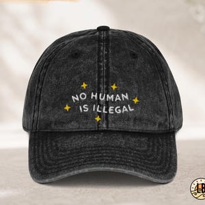 May include: A black acid-wash baseball cap featuring the embroidered white text NO HUMAN IS ILLEGAL centred on the front panel. The text is surrounded by small, yellow, four-pointed star icons stitched into the fabric above and below the words.