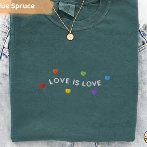 Könnte beinhalten: Ein blaugrünes T-Shirt mit der gestickten Aufschrift "LOVE IS LOVE" und bunten Herzmotiven. Eine goldene Halskette mit Anhänger liegt auf dem Shirt. Das T-Shirt ist auf einer Jeans gefaltet.
