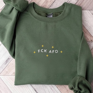 Könnte beinhalten: Ein olivgrüner Rundhals-Pullover mit weiß gesticktem Text FCK AFD, umgeben von kleinen gelben, vierzackigen Sternen. Das Kleidungsstück hat lange Ärmel mit gerippten Bündchen und einen gerippten Halsausschnitt.