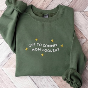 Könnte beinhalten: Ein olivgrüner Rundhals-Pullover mit weißem Sticktext, der OFF TO COMMIT MOM FOOLERY lautet, akzentuiert durch kleine gelbe, vierzackige Sticksterne, die den Schriftzug auf der Vorderseite des Kleidungsstücks umgeben.