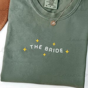Può includere: Una maglietta verde muschio con la scritta ricamata "THE BRIDE" e dettagli a stella gialla. È visibile una collana d'oro con un ciondolo. La maglietta è piegata e ha un'etichetta Comfort Colors.