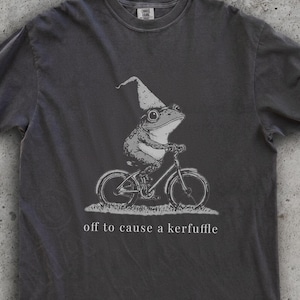 Könnte beinhalten: Dunkelgraues T-Shirt mit einer Illustration eines Frosches mit Zaubererhut auf einem Fahrrad. Der Text darunter lautet "off to cause a kerfuffle". Das Shirt hat einen Rundhalsausschnitt und kurze Ärmel.