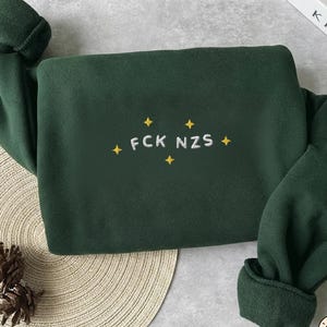 Op de afbeelding: Een donker bosgroen sweatshirt met lange mouwen en wit geborduurde tekst FCK NZS, omringd door kleine gele vierpuntige sterren. Het kledingstuk heeft een ronde hals en geribbelde boorden aan de mouwen.