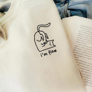 Könnte beinhalten: Cremefarbenes Sweatshirt mit einer schwarzen Grafik einer Katze und einer kleineren Katze, darunter der Text "i'm fine". Das Sweatshirt ist gefaltet, mit einem Buch und Jeans im Hintergrund.