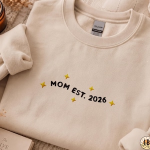 Puede incluir: Una sudadera de cuello redondo en color beige con texto bordado en negro que dice MOM EST. 2026, acentuado por pequeñas estrellas amarillas de cuatro puntas bordadas alrededor de las letras. La prenda tiene un cuello acanalado y etiqueta Gildan.