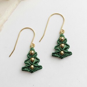 May include: A pair of Christmas tree earrings. Each earring features a green macrame tree design with three gold-colored beads. The earrings have gold-colored ear wires and are displayed on a white surface.