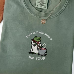 May include: An olive green t-shirt featuring the embroidered text "That's it, You're going in the SOUP." The design shows a frog chef stirring a pot. The shirt has a crew neck and a small tag.