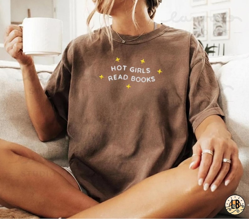 K&ouml;nnte beinhalten: Ein braunes T-Shirt mit dem Text "HOT GIRLS READ BOOKS" in Wei&szlig; und gelben Sternen. Das l&auml;ssige T-Shirt ist f&uuml;r den Alltag geeignet. Die Person h&auml;lt einen wei&szlig;en Becher.