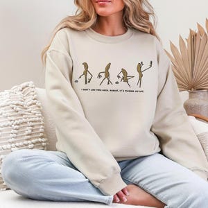 May include: Beige sweatshirt with a humorous graphic of stick figures interacting with rocks, accompanied by the text "I don't like this rock, Robert, it's pissing me off." The sweatshirt is a neutral color.