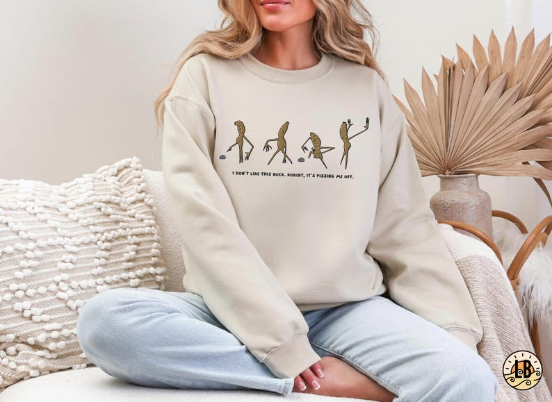 May include: Beige sweatshirt with a humorous graphic of stick figures interacting with rocks, accompanied by the text "I don't like this rock, Robert, it's pissing me off." The sweatshirt is a neutral color.