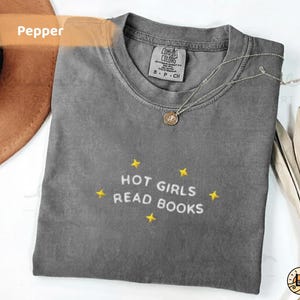 K&ouml;nnte beinhalten: Graues T-Shirt in Pfefferfarbe mit dem gestickten Text "HOT GIRLS READ BOOKS" in Wei&szlig;, umgeben von gelben Sternen. Eine silberne Halskette mit Anh&auml;nger liegt auf dem Shirt.