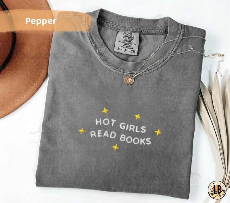 K&ouml;nnte beinhalten: Graues T-Shirt in Pfefferfarbe mit dem gestickten Text "HOT GIRLS READ BOOKS" in Wei&szlig;, umgeben von gelben Sternen. Eine silberne Halskette mit Anh&auml;nger liegt auf dem Shirt.