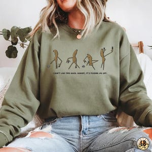 May include: Olive green sweatshirt with a humorous cartoon graphic of four figures. The text below the graphic reads, "I DON'T LIKE THIS ROCK. ROBERT, IT'S PISSING ME OFF."