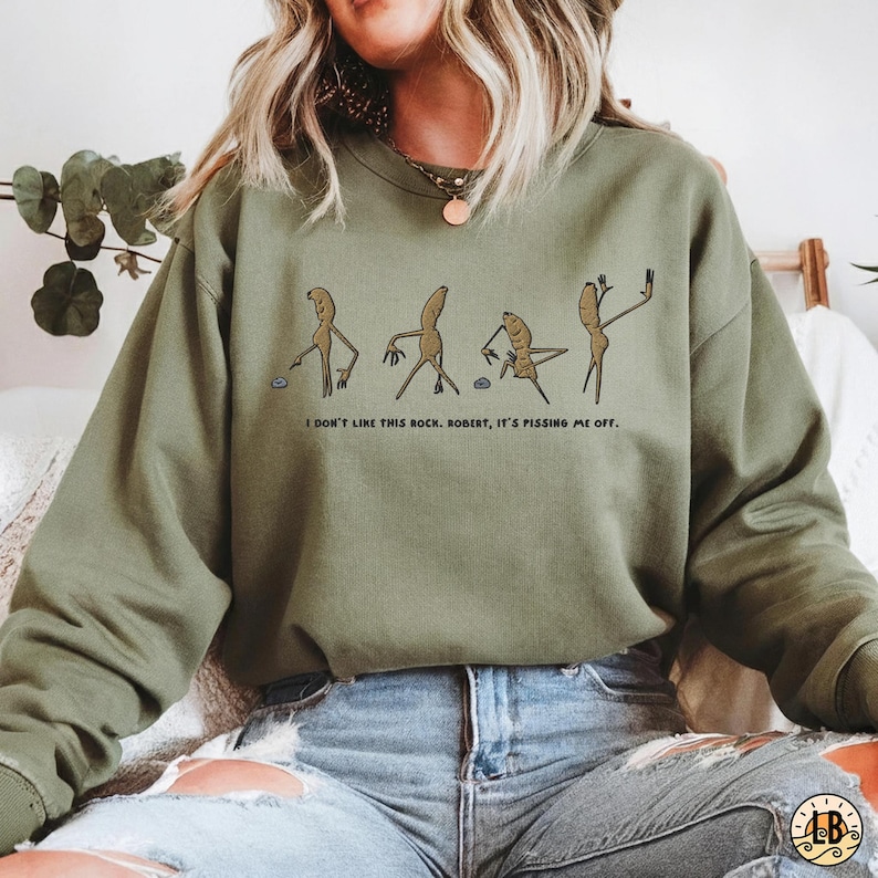 May include: Olive green sweatshirt with a humorous cartoon graphic of four figures. The text below the graphic reads, "I DON'T LIKE THIS ROCK. ROBERT, IT'S PISSING ME OFF."
