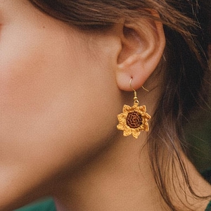 Può includere: Primo piano di un orecchino a forma di girasole all'uncinetto con un gancio color oro. L'orecchino presenta uno strato esterno giallo e un centro marrone, che imita l'aspetto di un girasole. L'orecchino è indossato all'orecchio di una persona.
