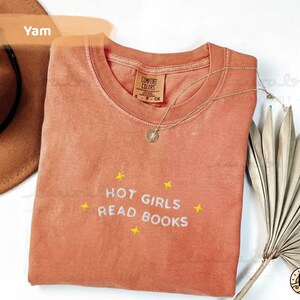 K&ouml;nnte beinhalten: Ein korallenfarbenes T-Shirt mit dem Text "HOT GIRLS READ BOOKS", der in Wei&szlig; mit gelben Sternen gestickt ist. Eine goldene Halskette mit Anh&auml;nger liegt auf dem Shirt. Das Shirt ist von Comfort Colors.