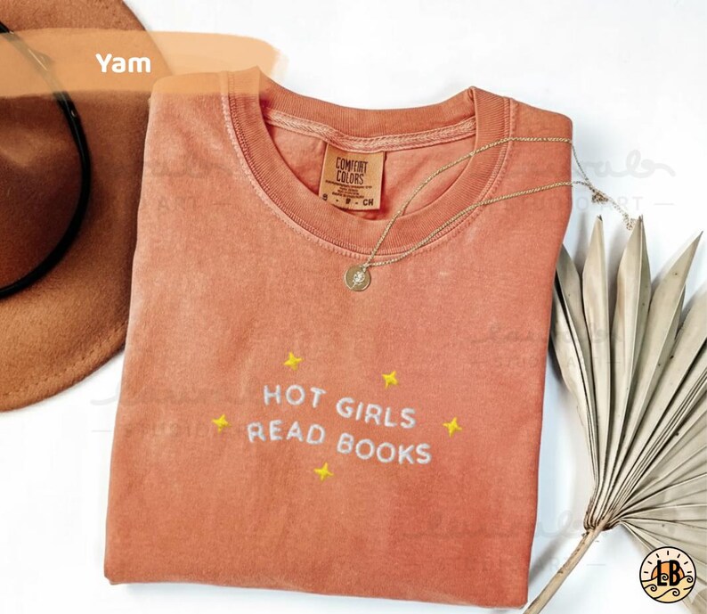 K&ouml;nnte beinhalten: Ein korallenfarbenes T-Shirt mit dem Text "HOT GIRLS READ BOOKS", der in Wei&szlig; mit gelben Sternen gestickt ist. Eine goldene Halskette mit Anh&auml;nger liegt auf dem Shirt. Das Shirt ist von Comfort Colors.