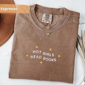 K&ouml;nnte beinhalten: Espressofarbenes T-Shirt mit dem gestickten Schriftzug "HOT GIRLS READ BOOKS" in Wei&szlig;, akzentuiert mit gelben Sternen. Eine silberne Halskette mit Anh&auml;nger liegt auf dem Shirt. Das Shirt ist von der Marke Comfort Colors, Gr&ouml;&szlig;e S-CH.