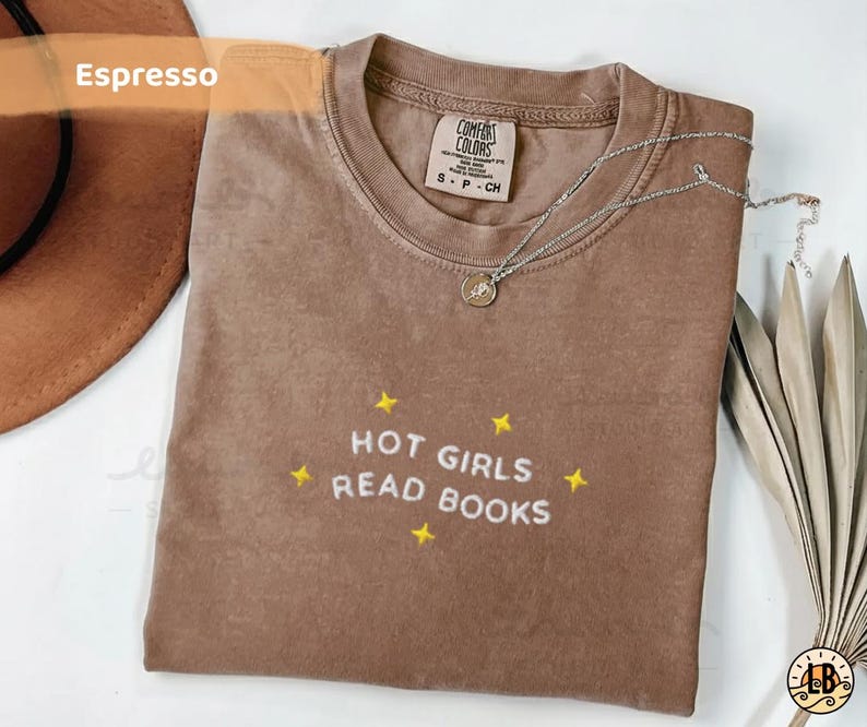 K&ouml;nnte beinhalten: Espressofarbenes T-Shirt mit dem gestickten Schriftzug "HOT GIRLS READ BOOKS" in Wei&szlig;, akzentuiert mit gelben Sternen. Eine silberne Halskette mit Anh&auml;nger liegt auf dem Shirt. Das Shirt ist von der Marke Comfort Colors, Gr&ouml;&szlig;e S-CH.