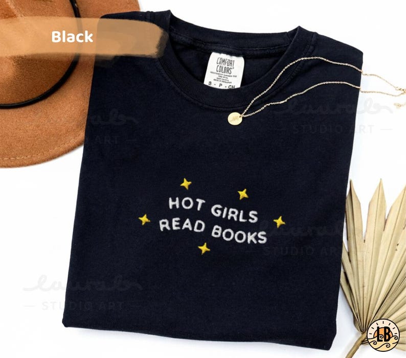 K&ouml;nnte beinhalten: Schwarzes Rundhals-T-Shirt mit dem gestickten Text "HOT GIRLS READ BOOKS" in Wei&szlig; und gelben Sternen. Eine goldene Halskette mit rundem Anh&auml;nger liegt &uuml;ber dem Shirt. Ein brauner Hut ist in der oberen linken Ecke zu sehen.