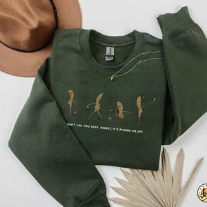 May include: A dark green sweatshirt with a crew neck and long sleeves. The front features an embroidered design of cartoon figures and the text "DON'T LIKE THIS ROCK, ROBERT, IT'S PISSING ME OFF."