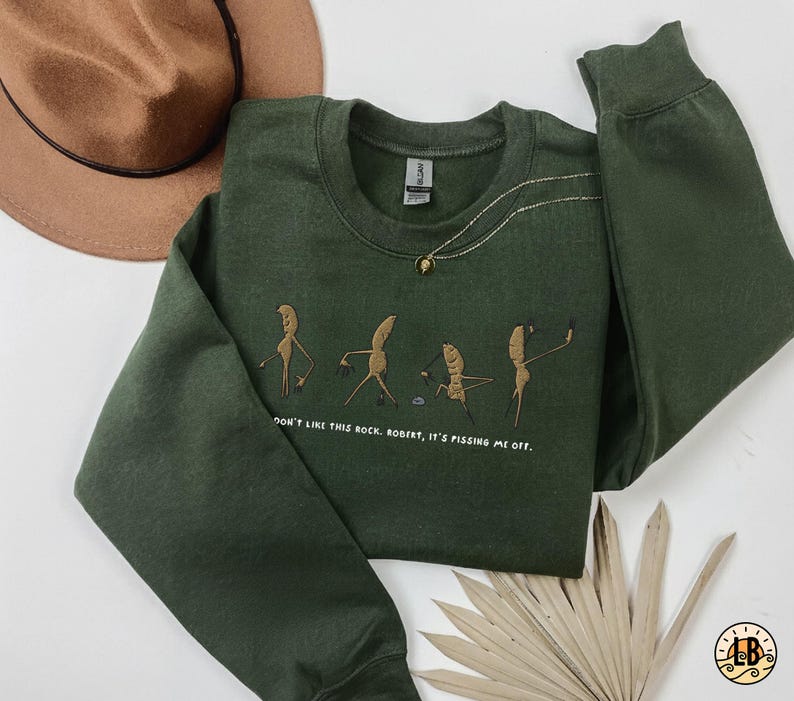May include: A dark green sweatshirt with a crew neck and long sleeves. The front features an embroidered design of cartoon figures and the text "DON'T LIKE THIS ROCK, ROBERT, IT'S PISSING ME OFF."