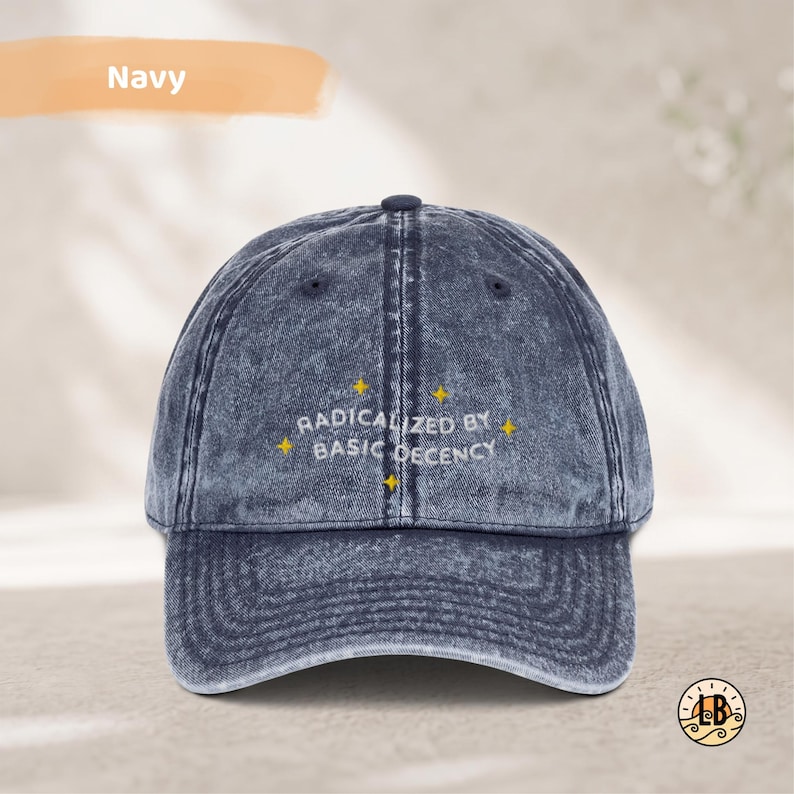 Op de afbeelding: Een marineblauwe baseballpet met een acid-wash effect, voorzien van de geborduurde witte tekst "RADICALIZED BY BASIC DECENCY" in het midden van het voorpaneel, geaccentueerd door kleine gele vierpuntige sterren rondom.