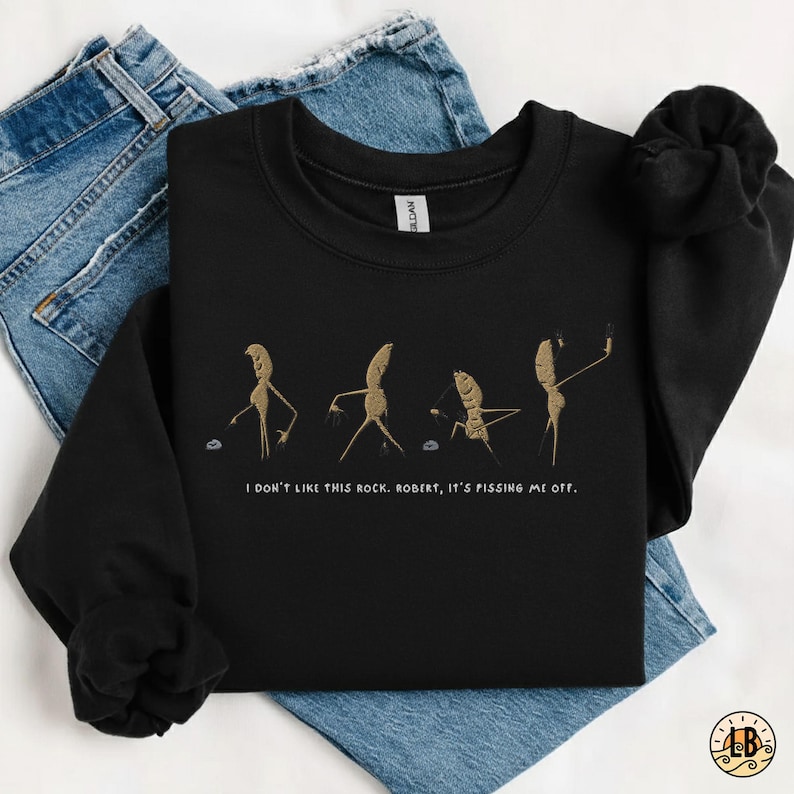May include: Black sweatshirt with a humorous graphic of anthropomorphic root vegetables in various poses, with the text "I don't like this rock. Robert, it's pissing me off." The sweatshirt is folded on top of a pair of blue jeans.