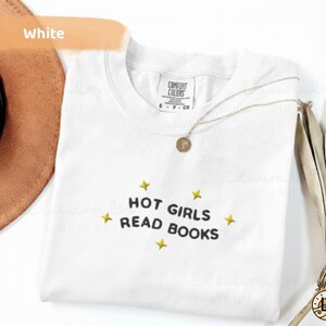 K&ouml;nnte beinhalten: Wei&szlig;es T-Shirt mit dem gestickten Text "HOT GIRLS READ BOOKS" in Schwarz, umgeben von gelben Sternen. Eine goldene Halskette und ein brauner Hut sind im Hintergrund zu sehen. Das Shirt ist von Comfort Colors.