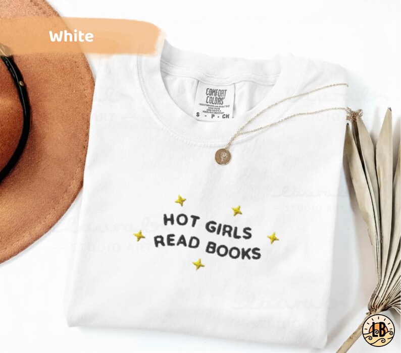 K&ouml;nnte beinhalten: Wei&szlig;es T-Shirt mit dem gestickten Text "HOT GIRLS READ BOOKS" in Schwarz, umgeben von gelben Sternen. Eine goldene Halskette und ein brauner Hut sind im Hintergrund zu sehen. Das Shirt ist von Comfort Colors.